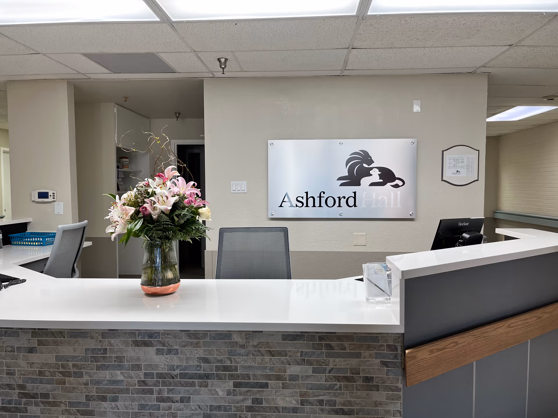 Reception desk area with a vase of pink and white flowers on a white countertop. Behind the desk is a sign on the wall that reads 'Ashford Hall' with a logo of a lion and a lamb. The area has a modern ceiling with recessed lighting and a tiled front on the desk.