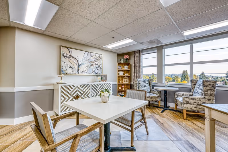 Bright communal seating area with tables and chairs, a decorative cabinet and bookshelf, and large windows overlooking trees.
