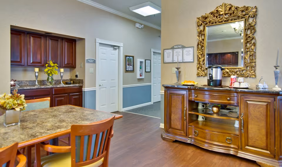 Interior view of a senior living facility dining area with a wooden table and chairs in the foreground. To the right, there is a wooden sideboard with a coffee maker, cups, and a decorative gold-framed mirror above it. On the left, there are dark wooden cabinets with a vase of yellow flowers and candles. The walls are beige with white trim, and there are doors and framed pictures in the background.