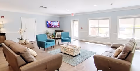 A bright and spacious living room in an assisted living facility with two brown sofas, two teal armchairs, a patterned ottoman, a light blue area rug, and a wall-mounted TV. Large windows allow natural light to fill the room.