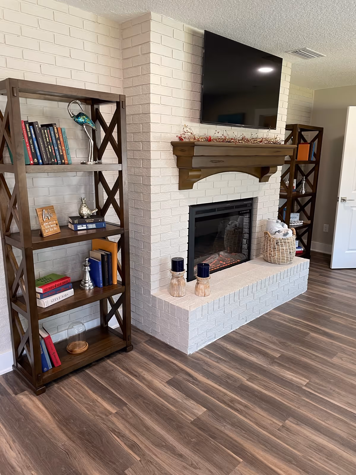 A cozy living room area featuring a white brick fireplace with a wooden mantel decorated with a garland. Above the mantel is a mounted flat-screen TV. On either side of the fireplace are dark wooden bookshelves filled with books and decorative items. The floor is covered with wood-patterned laminate flooring, and a basket with blankets is placed on the hearth.