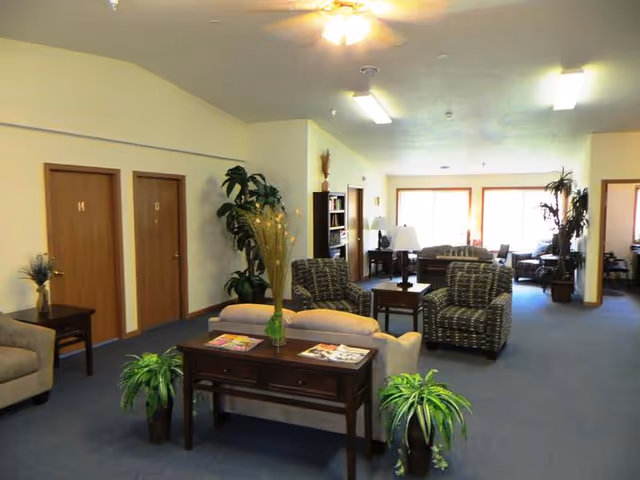 A spacious common area in a senior living facility with multiple seating arrangements including sofas and armchairs. The room has blue carpet, beige walls, several potted plants, a ceiling fan, and large windows letting in natural light. There are wooden doors along one wall and tables with magazines and decorative items.