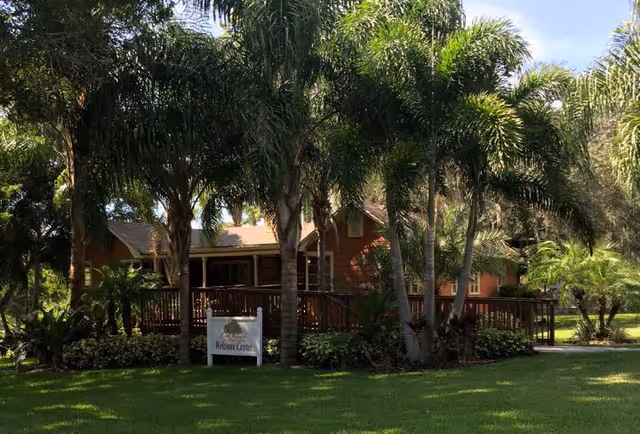 A wooden single-story building with a front deck surrounded by palm trees and a lawn, with a small 'Welcome Center' sign in front.