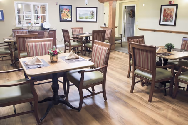 A dining room in a senior living facility with several wooden tables and chairs. Each table is set with placemats, folded napkins, and small floral centerpieces. The room has wood flooring, framed artwork on the walls, and a sideboard with decorative items.