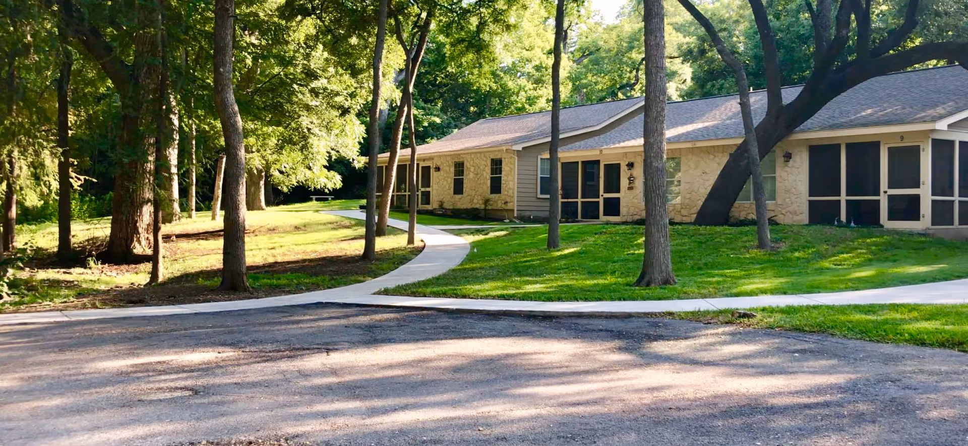 Single-story stone building with screened porches, a curving sidewalk, and trees on a sunlit lawn.