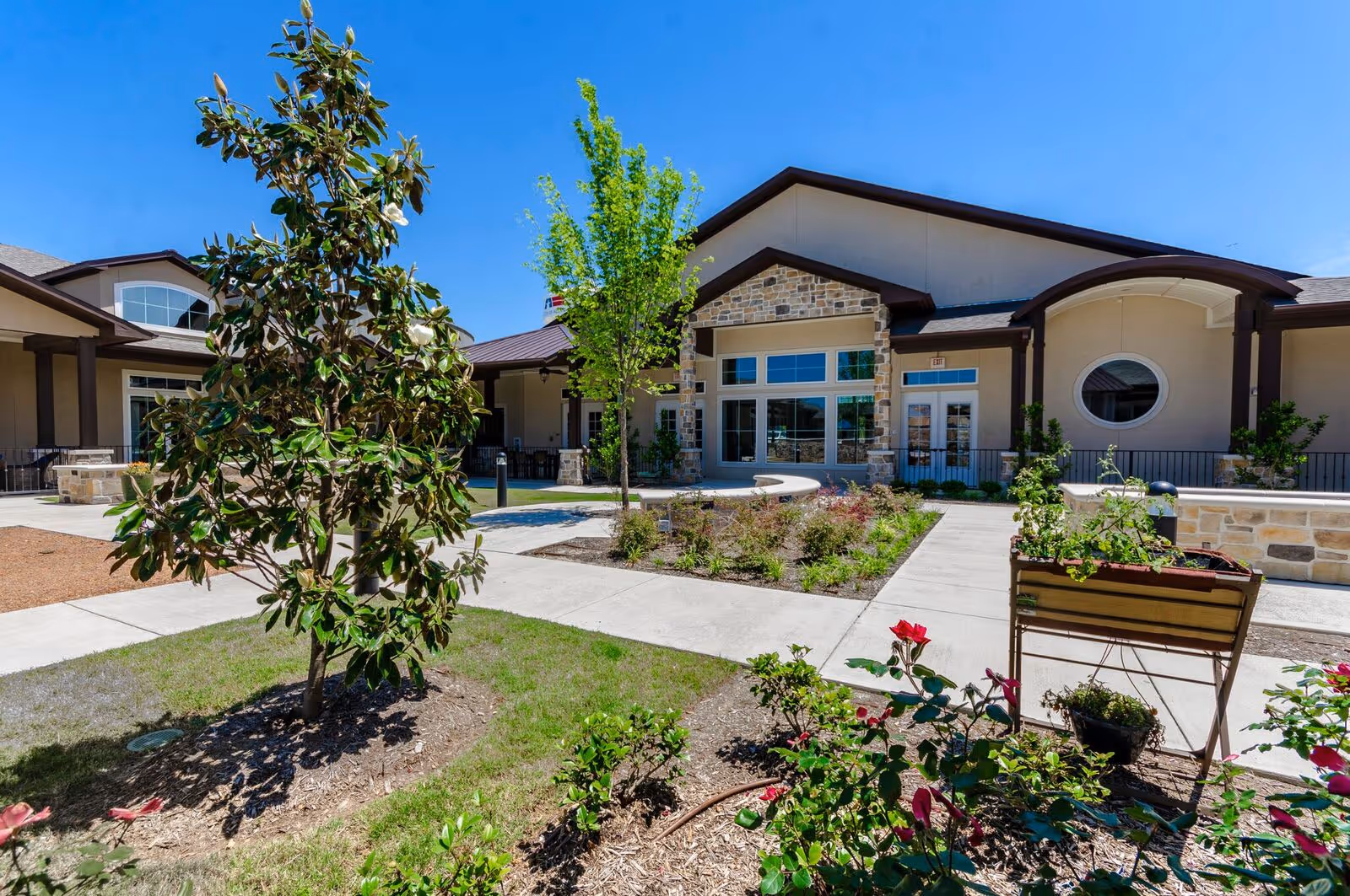 Outdoor courtyard area of a senior living facility with a clear blue sky, landscaped garden beds with small trees and flowering plants, concrete walkways, and a building with stone and beige exterior walls featuring large windows and a circular window.