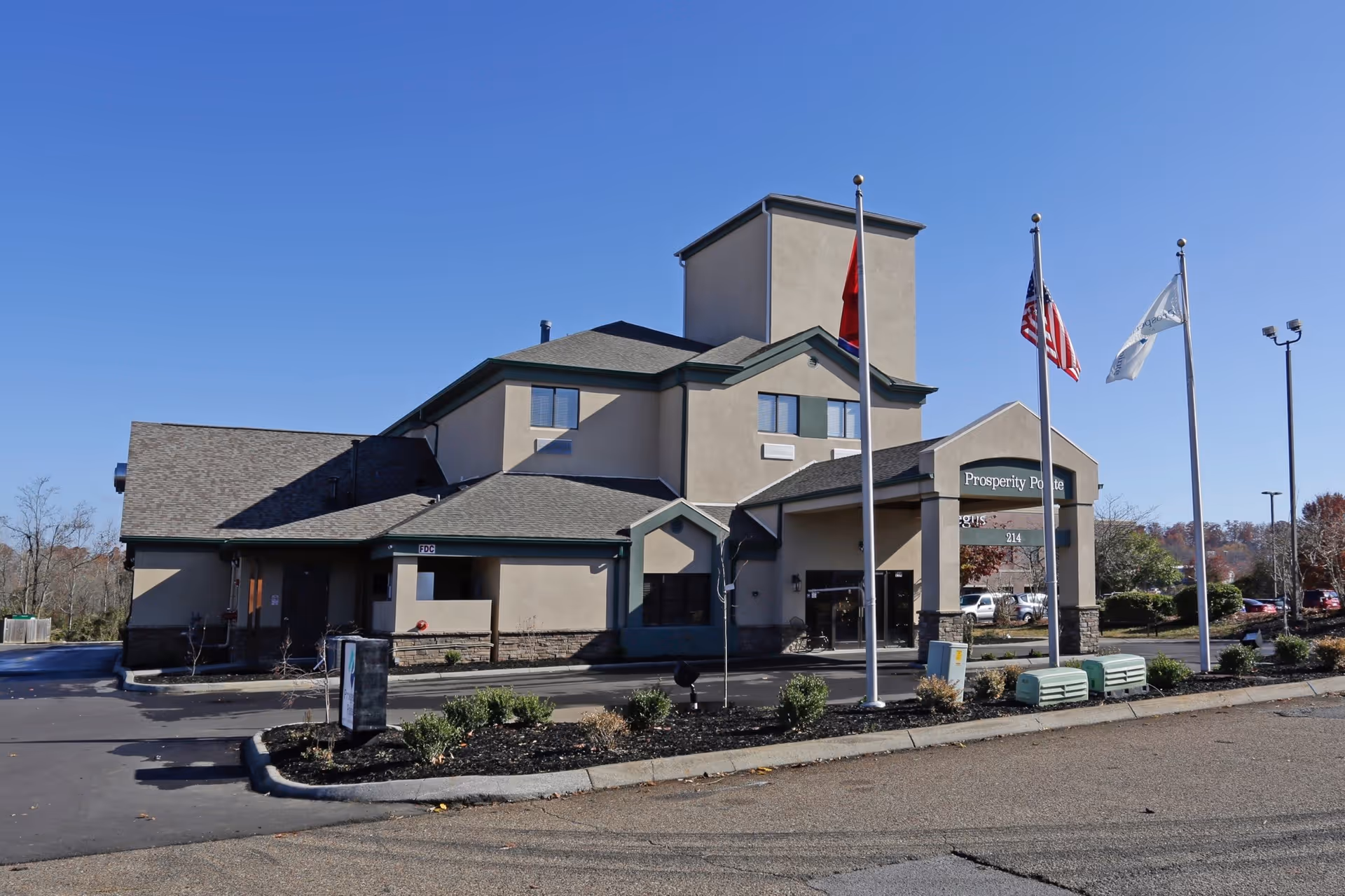 Exterior view of Prosperity Pointe facility building with beige walls and multiple roof levels under a clear blue sky. Three flagpoles stand in front, displaying a red flag, the American flag, and a white flag with text. There are some shrubs and a parking area around the building.