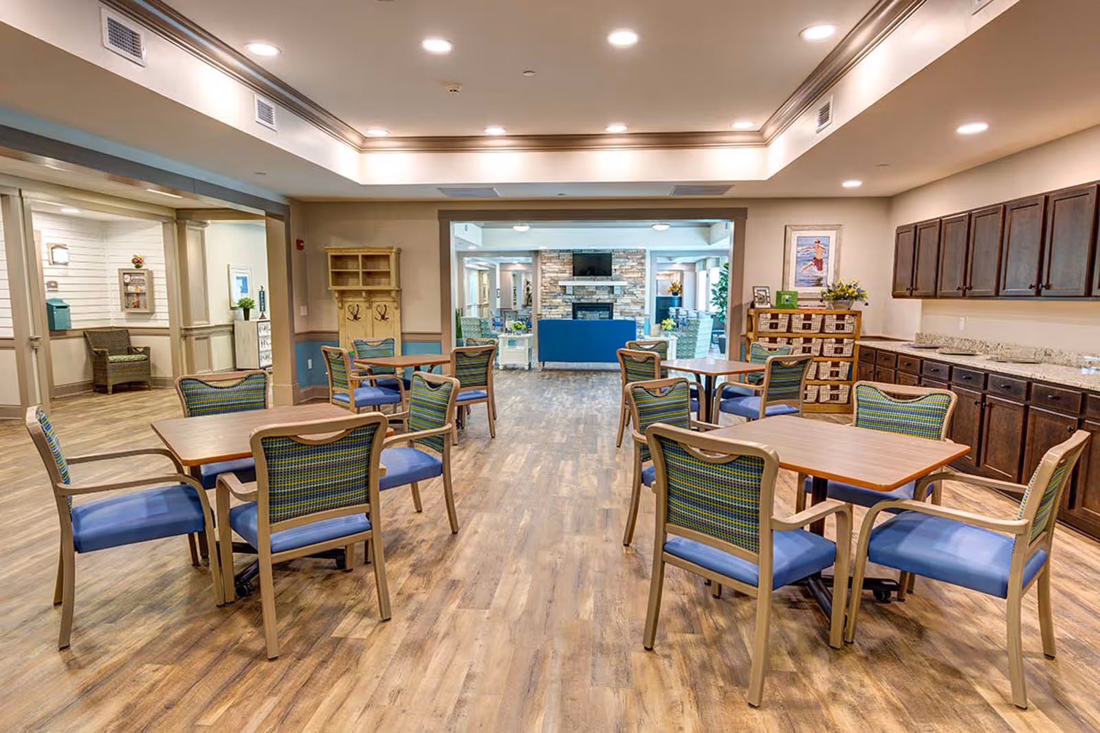 Spacious communal dining area with multiple wooden tables and blue-cushioned chairs, wood flooring and cabinetry.