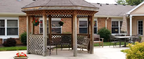 Wooden gazebo with benches and patio furniture on a concrete courtyard in front of a single-story brick senior living building with potted plants.