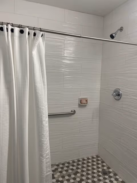 A shower area with white textured wall tiles and a white shower curtain hanging from a metal rod. There is a silver showerhead and a grab bar mounted on the wall. The floor has hexagonal tiles in shades of gray and white, and a soap holder with a bar of soap is attached to the wall.