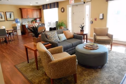 A bright and cozy living room area with a gray sofa adorned with patterned and solid pillows, two beige armchairs, and a round blue ottoman with a tray on top. The room features wooden flooring, a patterned area rug, a small wooden side table with a plant, and large windows letting in natural light. In the background, there is a kitchenette with wooden cabinets, a water cooler, and a dining table with chairs.