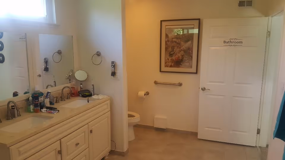 Accessible bathroom with a double-sink vanity, mirror, toilet, grab bar, and a door labeled "Bathroom".