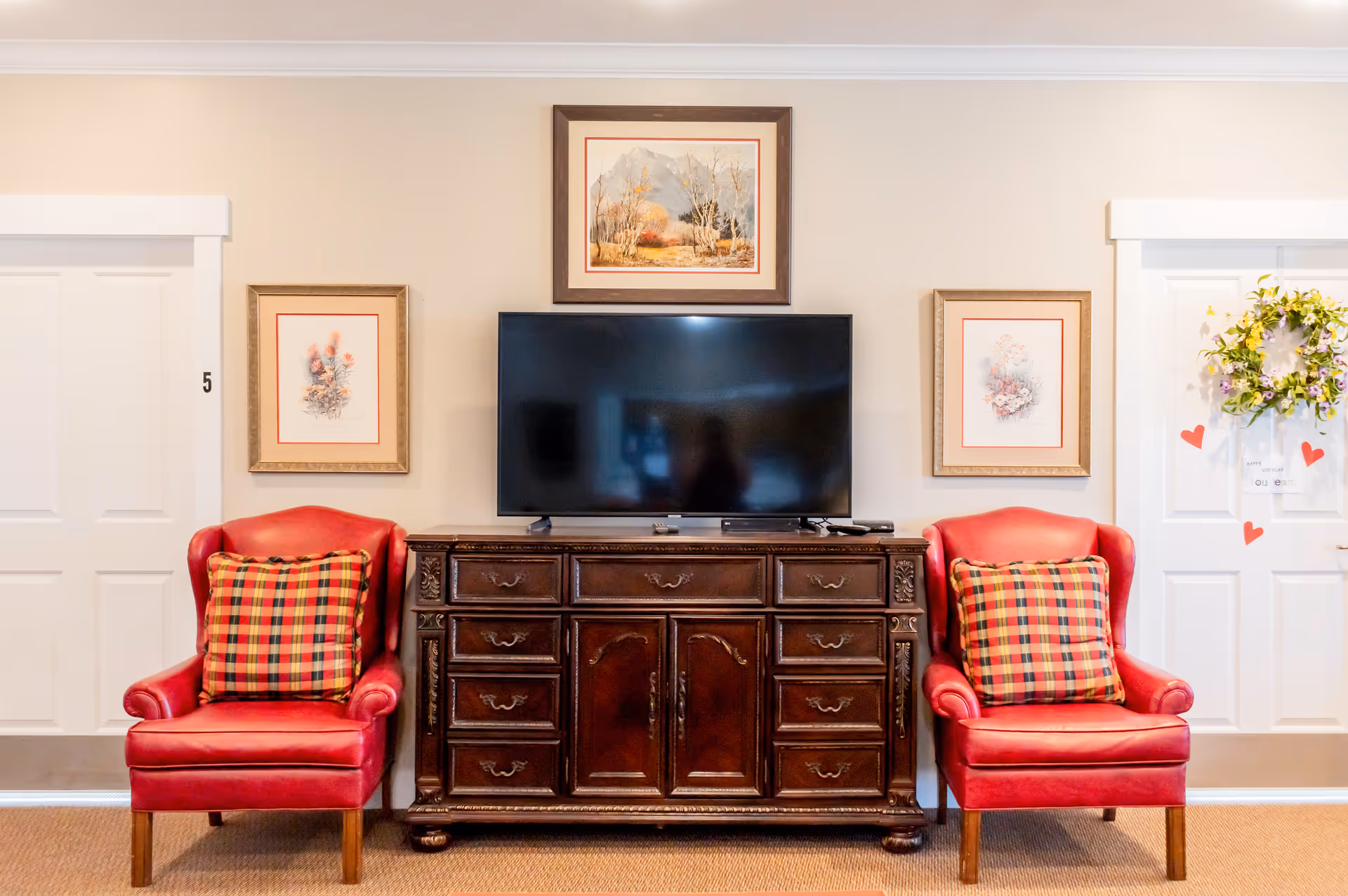 Two red armchairs with plaid pillows flank a dark wooden cabinet topped with a flat-screen TV and framed artwork on a beige wall.