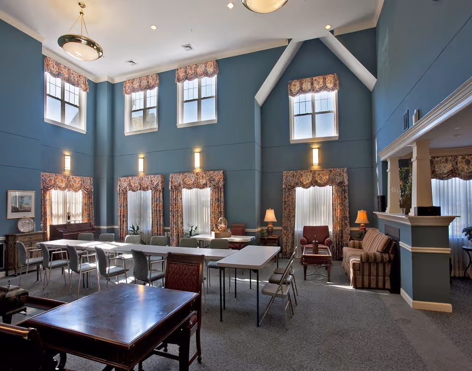 A spacious room with high ceilings painted in blue, featuring multiple windows with floral curtains. The room contains several tables and chairs arranged for group seating, along with a sitting area that includes armchairs, a sofa, and lamps. The room has a cozy and inviting atmosphere with traditional decor.