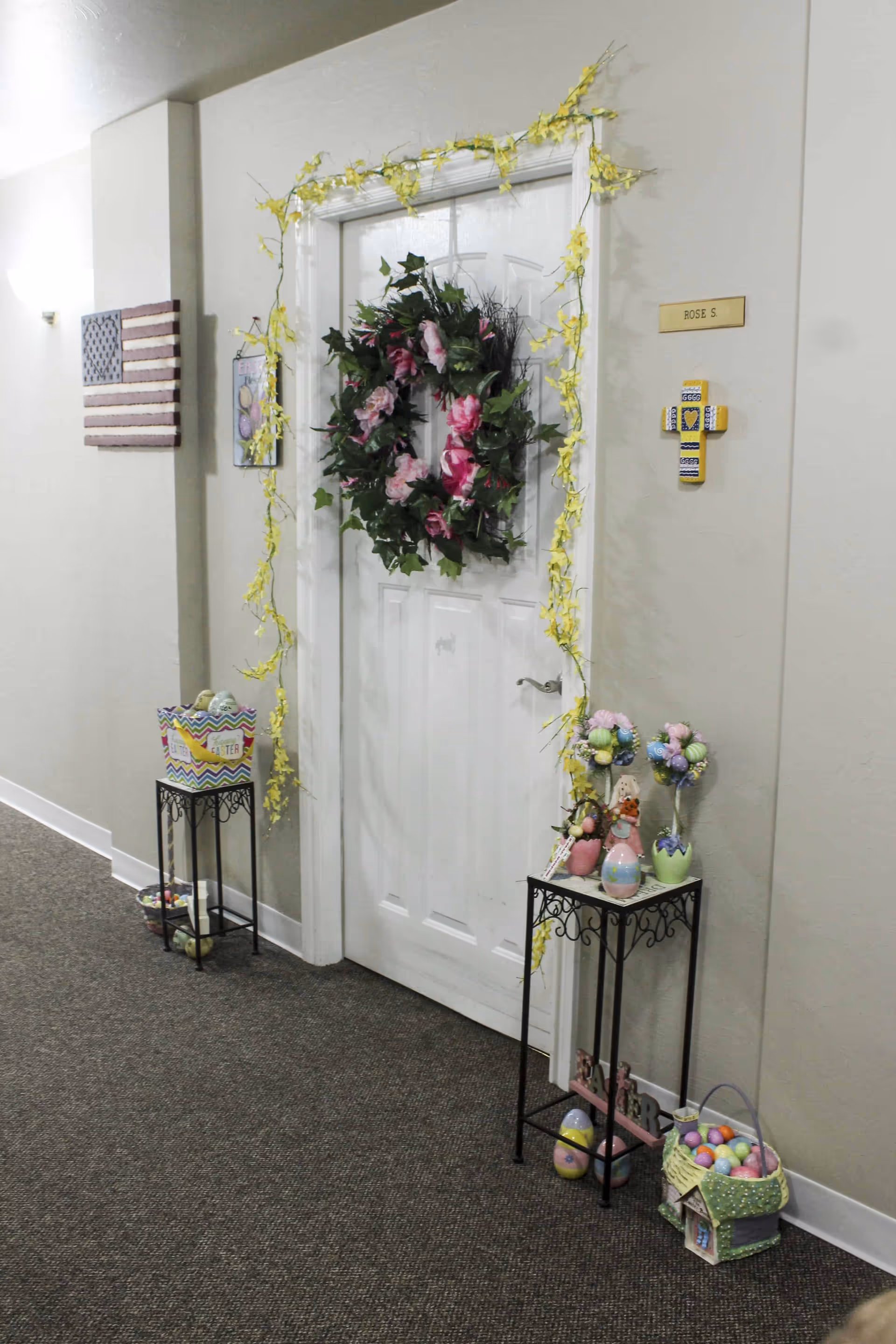 A white door decorated with a floral wreath and yellow garland in a hallway. The door has a nameplate that reads 'ROSE S.' and a decorative cross mounted on the wall beside it. Two small black metal stands hold Easter-themed decorations including colorful eggs and flower arrangements. An American flag wall decoration is visible on the left wall.