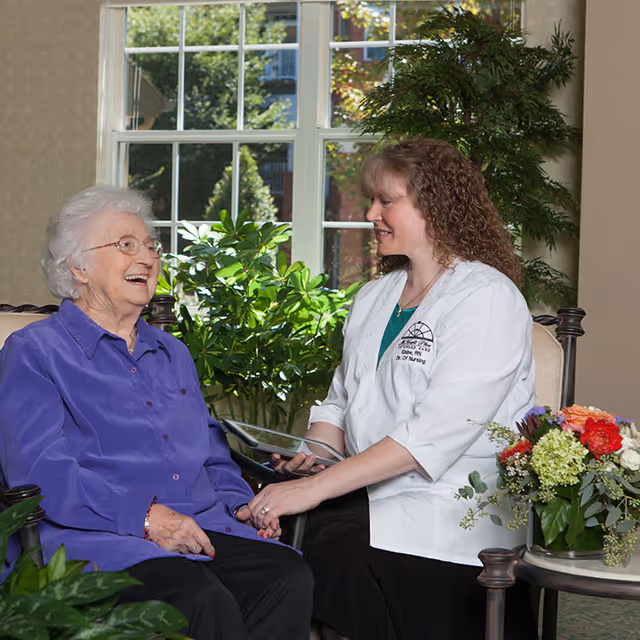 An elderly woman with white hair and glasses wearing a purple shirt is sitting and smiling while holding hands with a female healthcare professional in a white coat. They are seated in a well-lit room with large windows and green plants in the background. A table with a colorful flower arrangement is nearby.