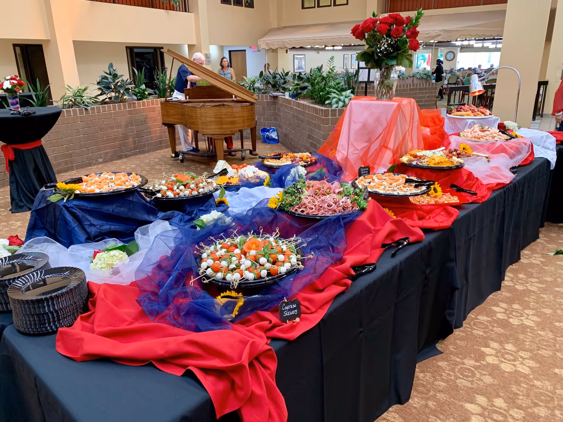 A buffet table set up with various appetizers including skewers, shrimp, and cheese platters, decorated with red, white, and blue cloths and sunflowers. In the background, there is a grand piano with a person playing it and another person standing nearby. The setting appears to be a spacious indoor common area with plants and seating.