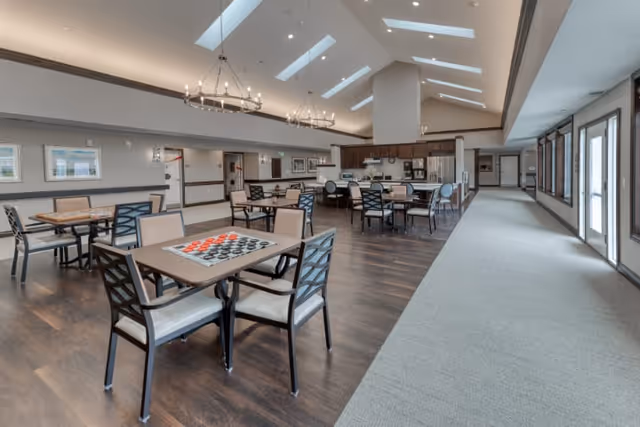 A spacious, well-lit common area with multiple tables and chairs arranged for socializing or playing games. One table in the foreground has a checkers board set up. The room features high ceilings with skylights, modern chandeliers, and a combination of wood flooring and carpet. Large windows and glass doors line one side of the room, allowing natural light to fill the space. The back of the room includes a kitchen area with stainless steel appliances and additional seating.