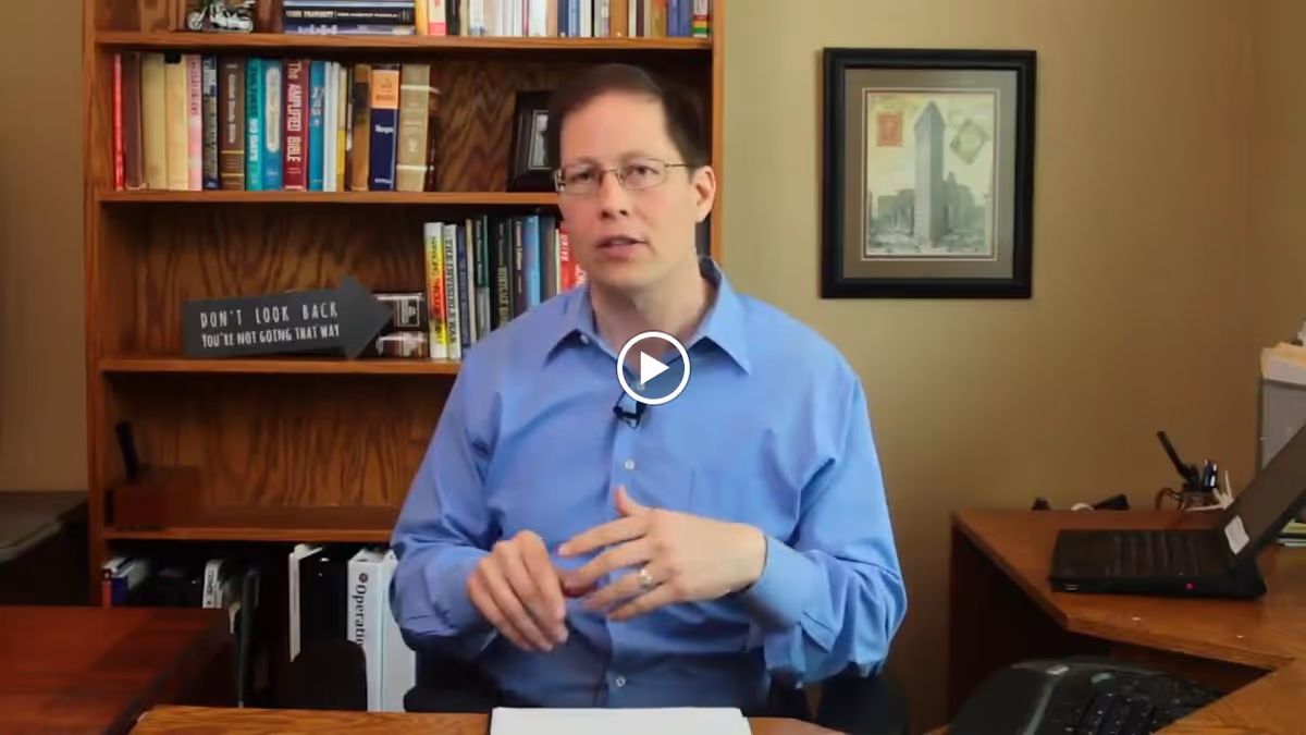 A man wearing glasses and a blue shirt is sitting at a desk in an office setting. Behind him is a wooden bookshelf filled with books and a decorative sign that reads 'DON'T LOOK BACK YOU'RE NOT GOING THAT WAY.' There is a framed picture on the wall and a laptop on a side desk.