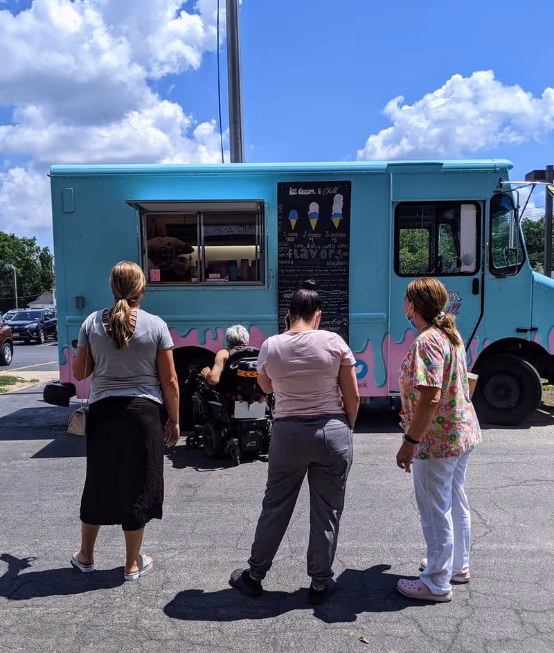 Four people standing in front of a turquoise and pink ice cream truck on a sunny day. One person is in a wheelchair near the service window of the truck, while the other three stand behind them. The sky is blue with scattered clouds.