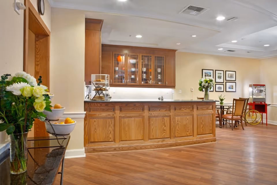 Interior view of a senior living facility's common area featuring a wooden reception or serving counter with cabinets and glass-front upper cupboards displaying dishes. The room has wood flooring, a small round table with chairs, a popcorn machine on a red cart, and framed pictures on the wall. A vase with flowers and a tiered fruit bowl are also visible.