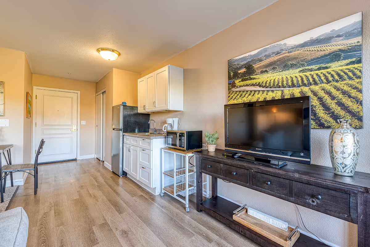 Interior view of a senior living facility room showing a small kitchenette with white cabinets, a stainless steel refrigerator, a microwave, and a coffee maker. Next to the kitchenette is a dark wooden console table with a flat-screen TV, a decorative vase, and a small potted plant. Above the TV is a large wall art depicting a vineyard landscape. The room has light wood flooring and beige walls, with a white door and closet in the background.