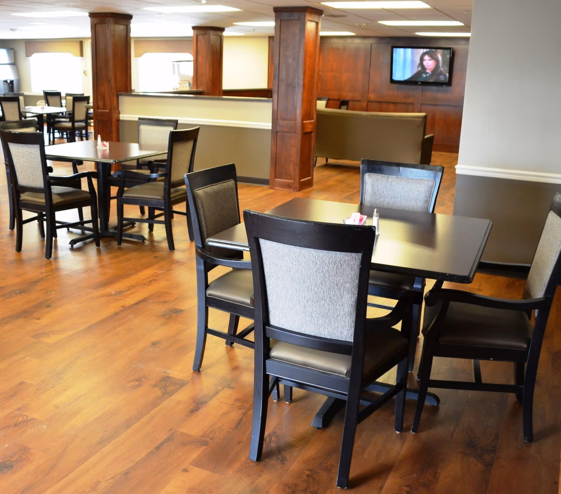A bright communal dining area with dark wood tables and upholstered chairs on a wood floor and a TV and seating area in the background.