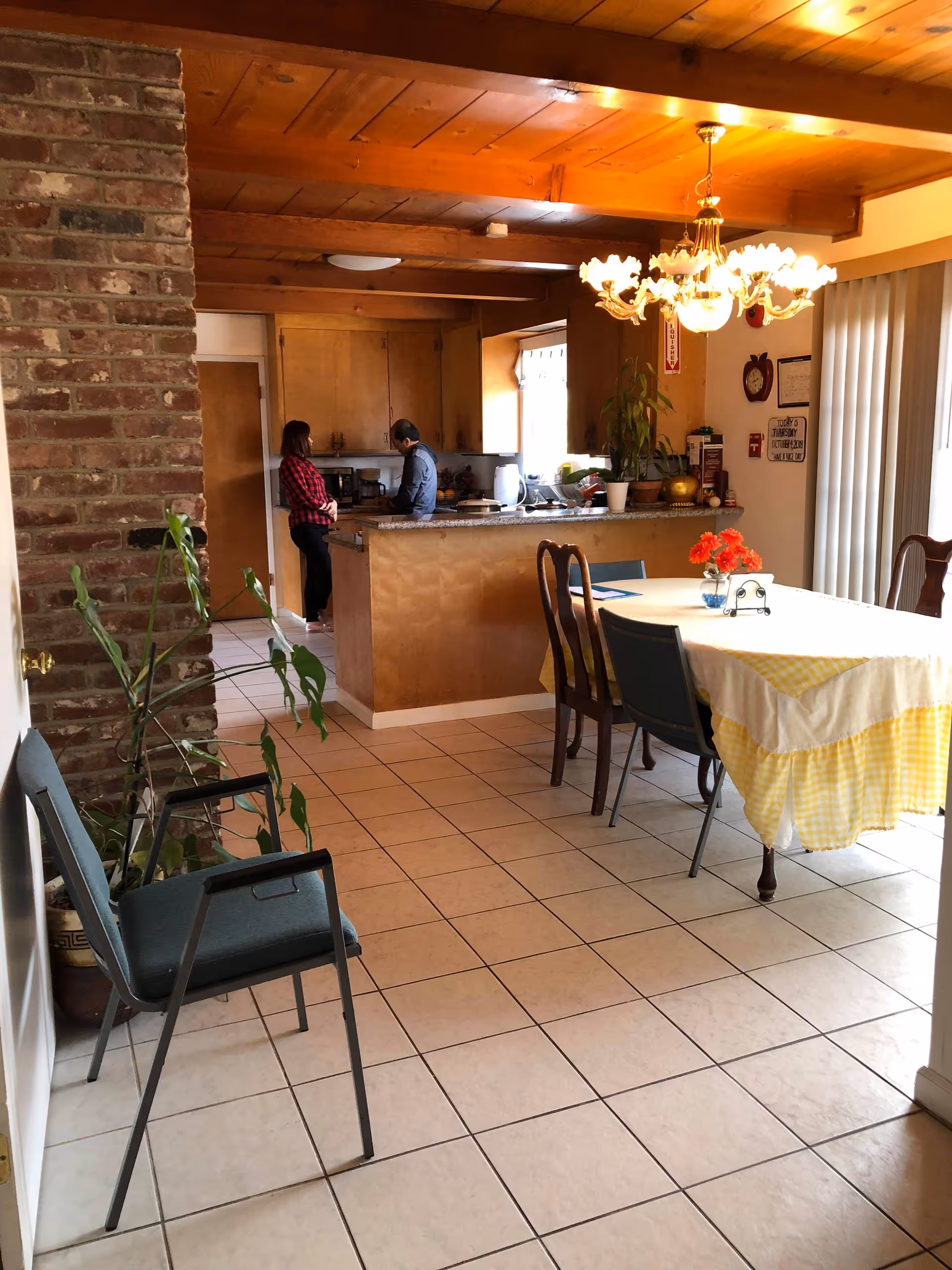 Interior view of a senior living facility dining area with a kitchen island, a chandelier, and two people in the background.