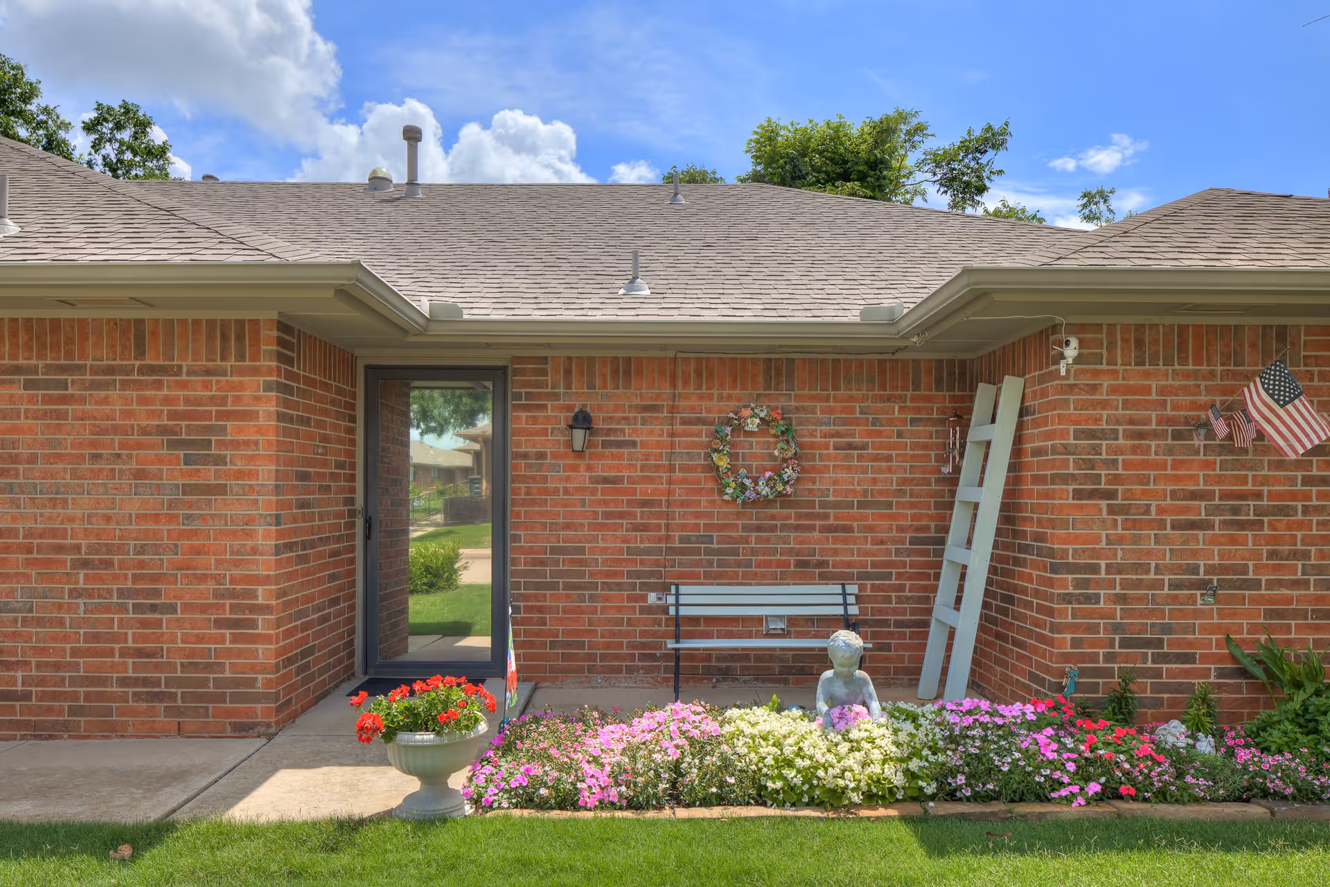 Exterior view of a brick building wall with a glass door, a bench, a decorative wreath, a white ladder leaning against the wall, and a flower bed with colorful flowers and a small statue of a child. An American flag is mounted on the wall, and the sky is partly cloudy.