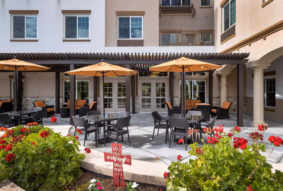 Outdoor courtyard patio of a senior living building with tables, chairs, orange umbrellas, lounge seating and red flowering plants in front of a multi-story facade.