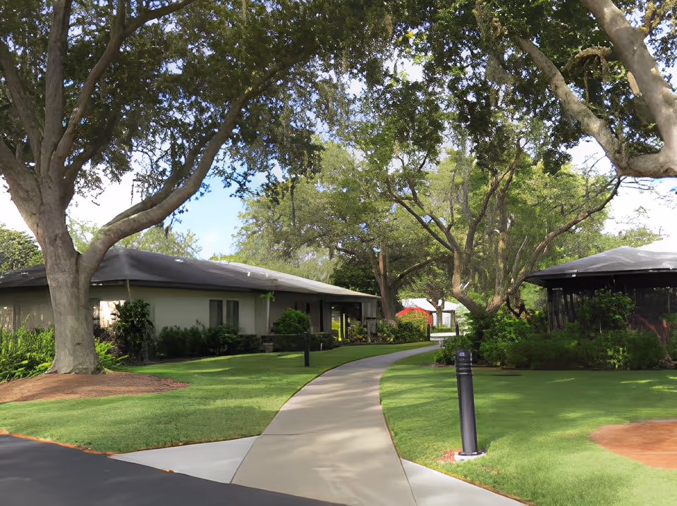 A paved walkway curves through a green lawn with large trees providing shade. Single-story buildings with dark roofs are visible on either side of the path, surrounded by bushes and greenery. The sky is clear and blue.