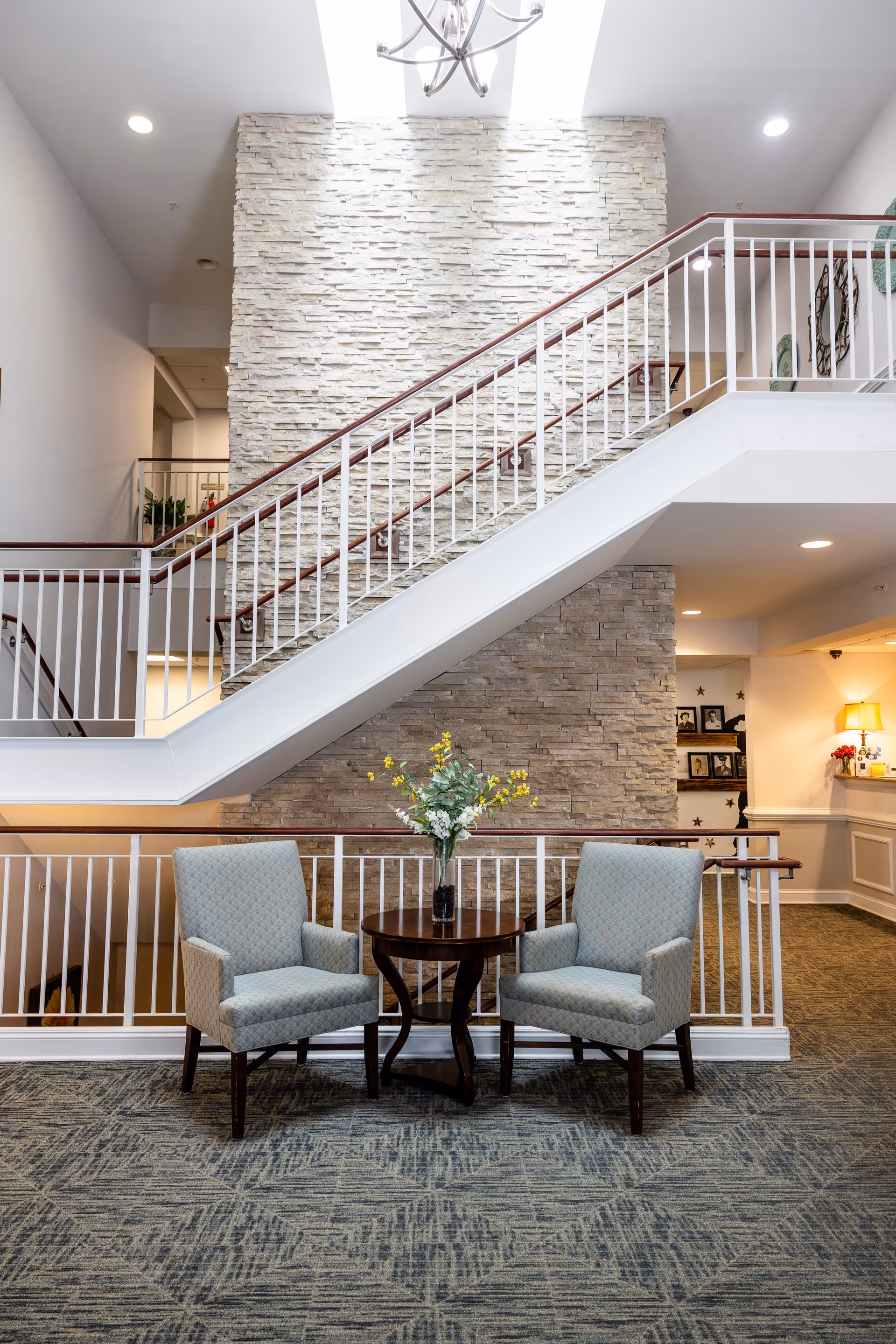Two upholstered chairs and a small table with a vase placed in front of an interior stairwell and stone accent wall in a senior living facility.