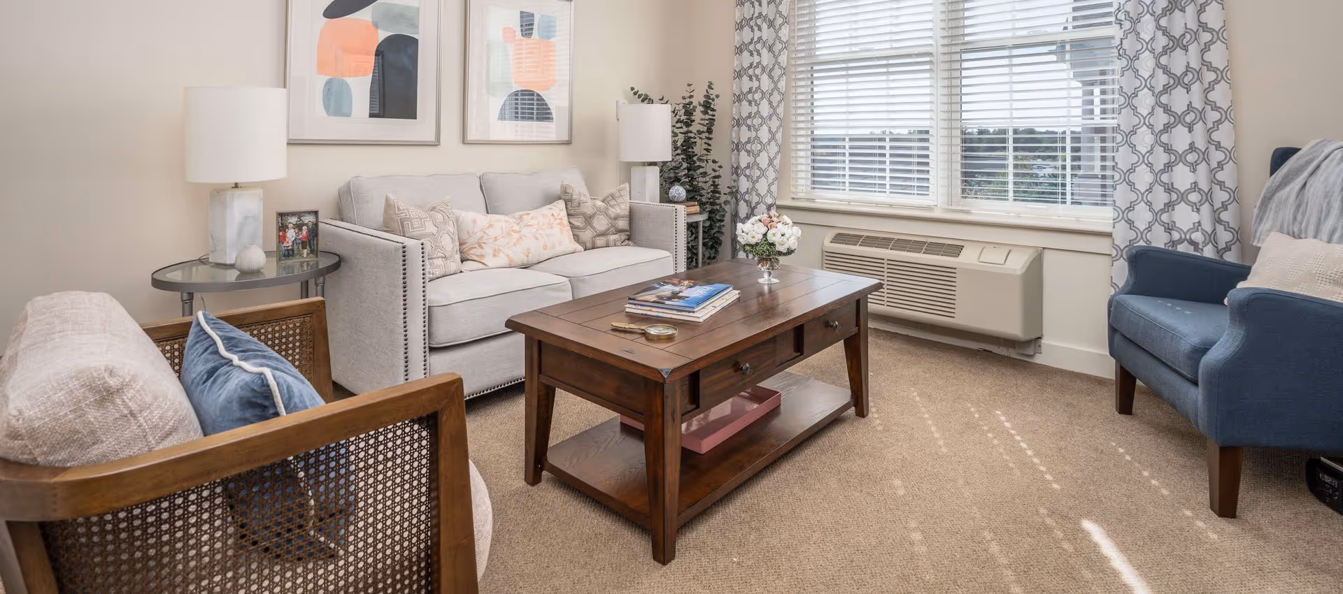 A cozy living room with a beige sofa adorned with patterned pillows, a wooden coffee table with books and a flower vase, a blue armchair with a pillow and throw blanket, a wooden chair with cushions, two table lamps on side tables, abstract wall art, and a window with patterned curtains and blinds.