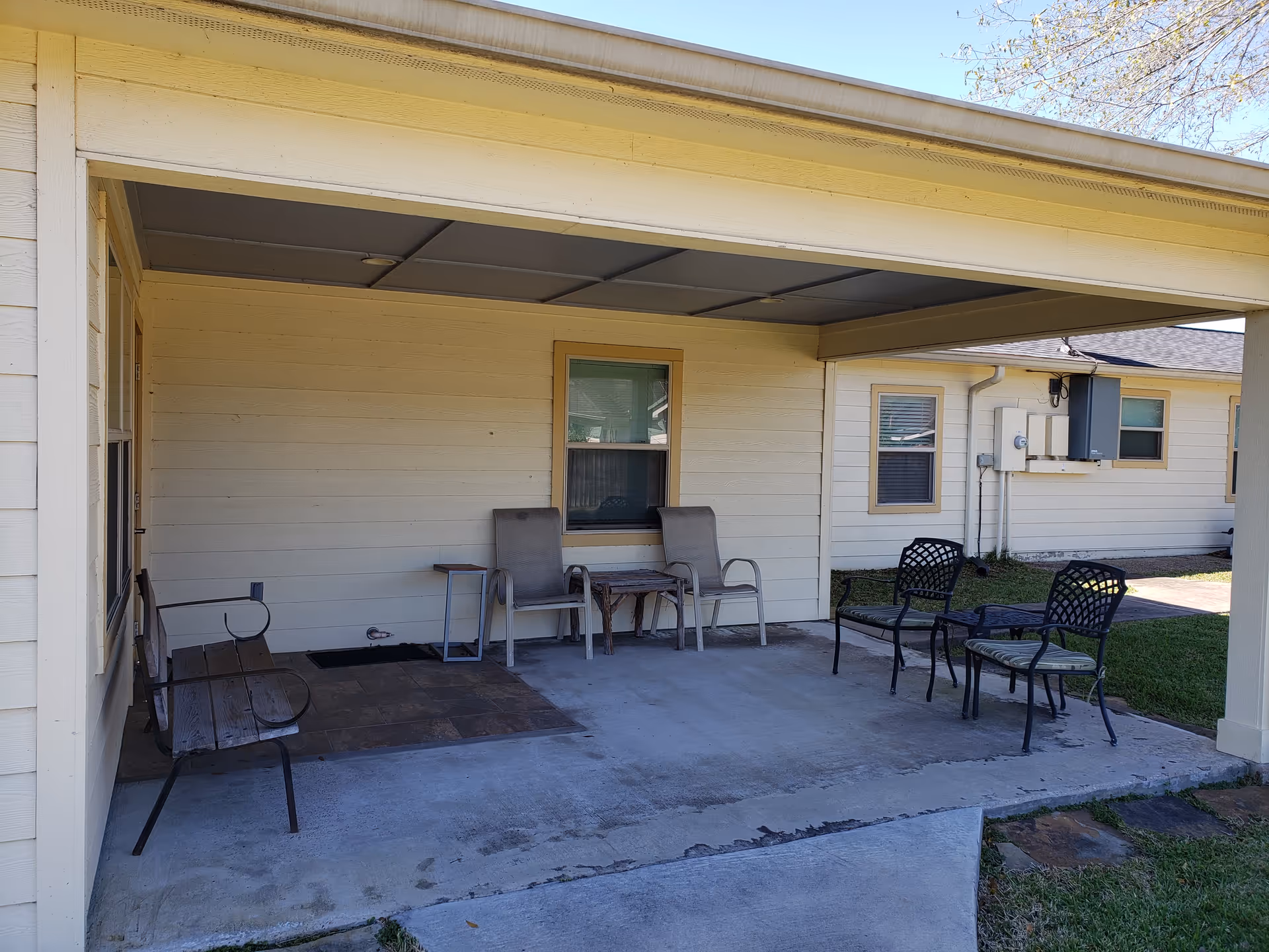 Covered outdoor patio area with several chairs and small tables arranged on a concrete floor, adjacent to a beige building with windows and a grassy lawn nearby.