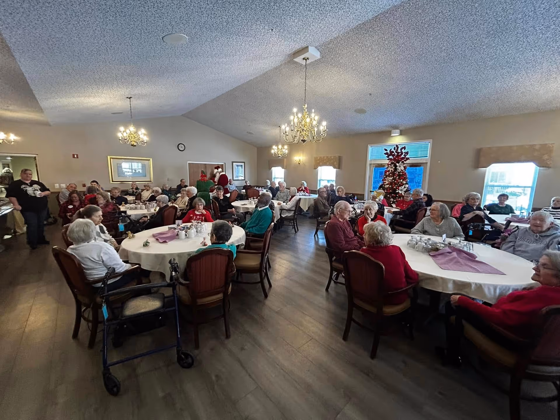 A large group of elderly people seated around multiple round tables in a spacious, well-lit dining room decorated with chandeliers and a Christmas tree. The room has beige walls, wooden flooring, and several windows letting in natural light. Some people are engaged in conversation, and a few staff members are present.