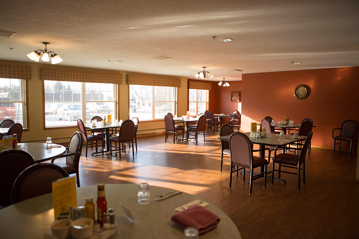 A dining room with several round and square tables, each surrounded by chairs. The room has large windows letting in natural light, with a reddish-brown accent wall featuring a clock. Tables are set with condiments, menus, and glassware.