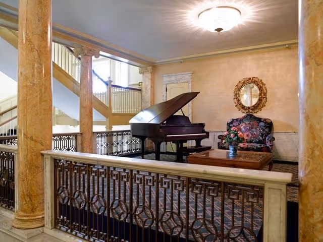 An elegant interior space featuring a grand piano, a floral upholstered armchair, a wooden coffee table with a flower vase, ornate gold-framed mirror on the wall, marble columns, and a staircase in the background.