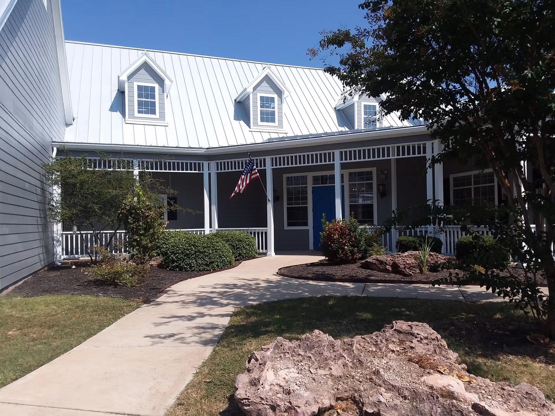 Front exterior view of a building with a white metal roof, gray siding, a blue door, and a porch with white railings. There is an American flag hanging near the entrance and landscaping with bushes, trees, and rocks along a concrete walkway leading to the door.