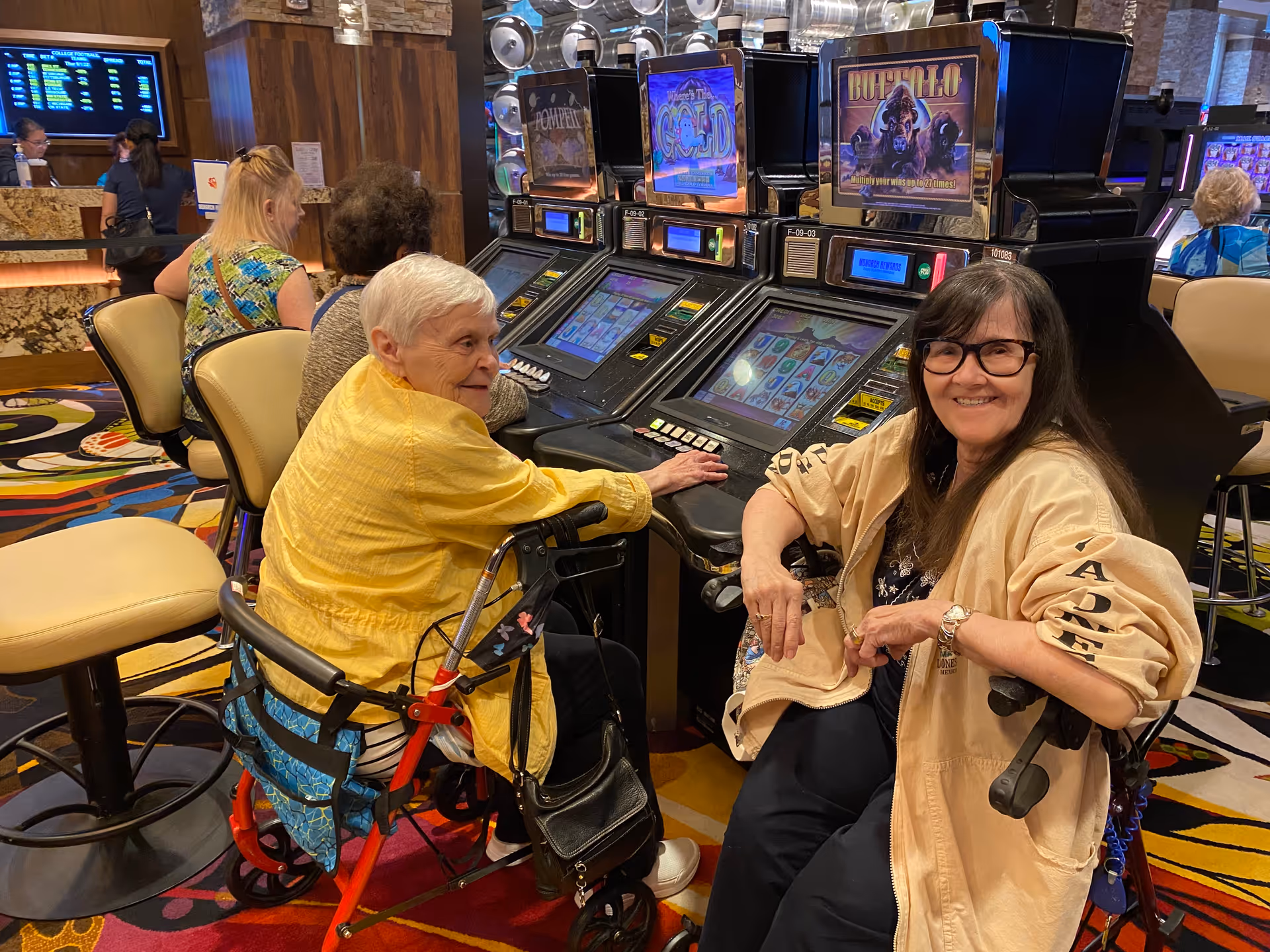 Two elderly women sitting and smiling near slot machines in a casino-like setting. One woman is using a walker and wearing a yellow jacket, while the other woman is wearing glasses and a beige jacket. Other people are visible in the background, also seated and engaged with the slot machines.
