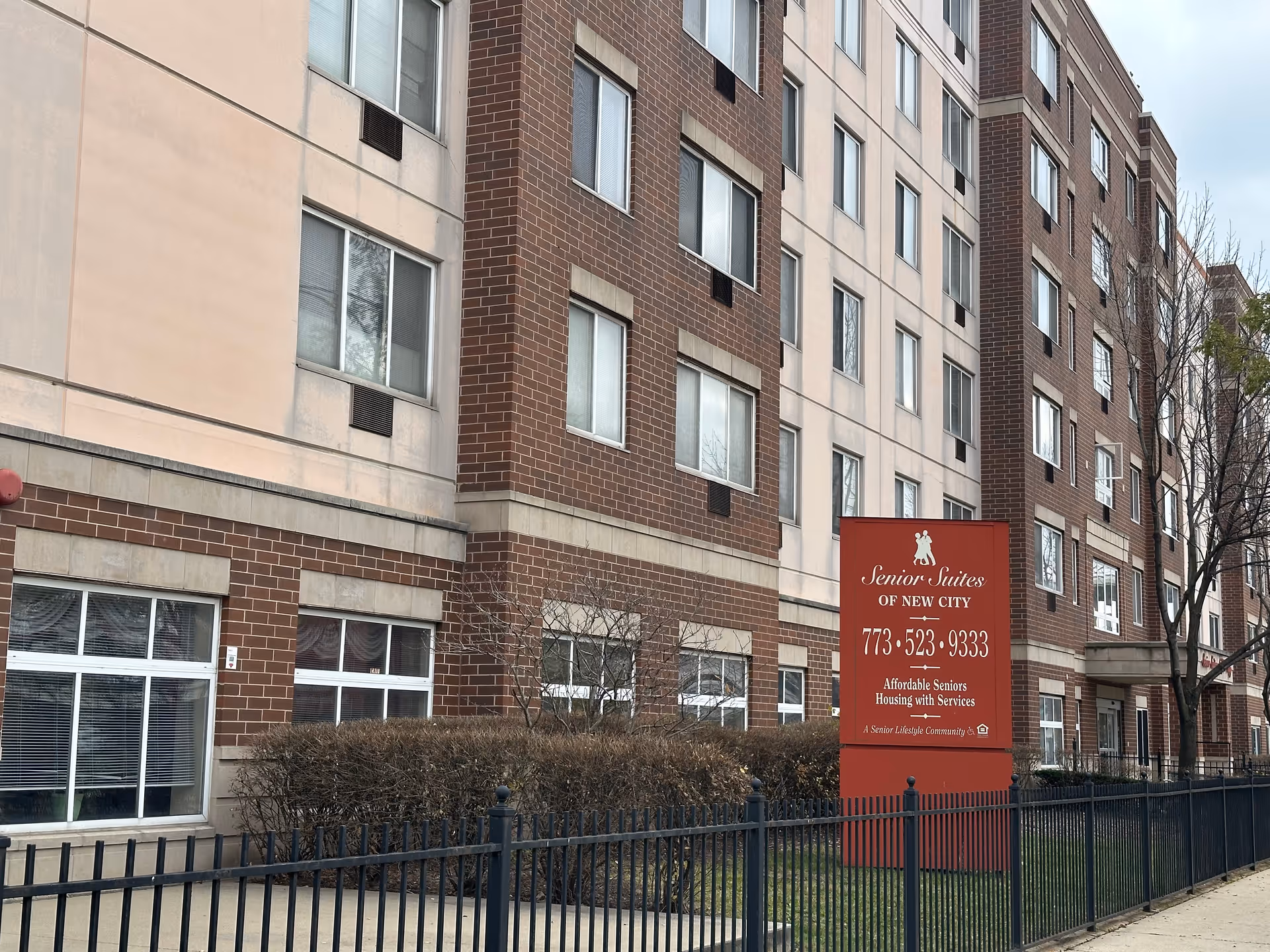 Exterior view of a multi-story brick-and-stucco senior living building with a red 'Senior Suites of New City' sign and a metal fence in front.