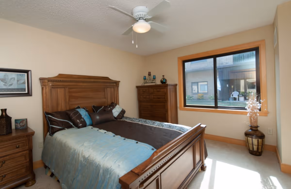 A bedroom with a wooden bed frame and headboard, made up with a blue and brown bedspread and multiple pillows. There is a wooden nightstand with decorative items on the left side of the bed, a wooden dresser against the far wall, and a large window with a view of an outdoor area. A ceiling fan with a light is mounted on the ceiling, and a decorative vase with dried branches is placed near the window.
