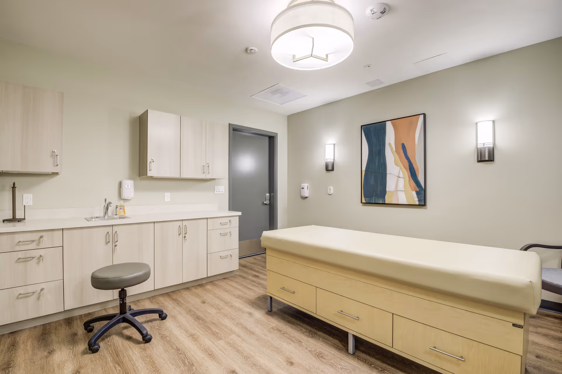 A clean and modern medical examination room with a padded examination table, a rolling stool, light wood cabinets with a sink, a gray door, abstract wall art, and two wall-mounted lights.