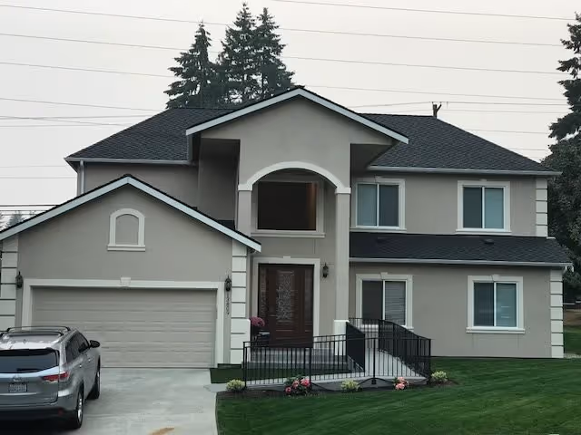 Front exterior view of a two-story residential building with a gray roof, beige walls, a double garage, a ramp leading to the front door, and a silver SUV parked in the driveway. There are some small plants and flowers near the entrance and green grass surrounding the house.