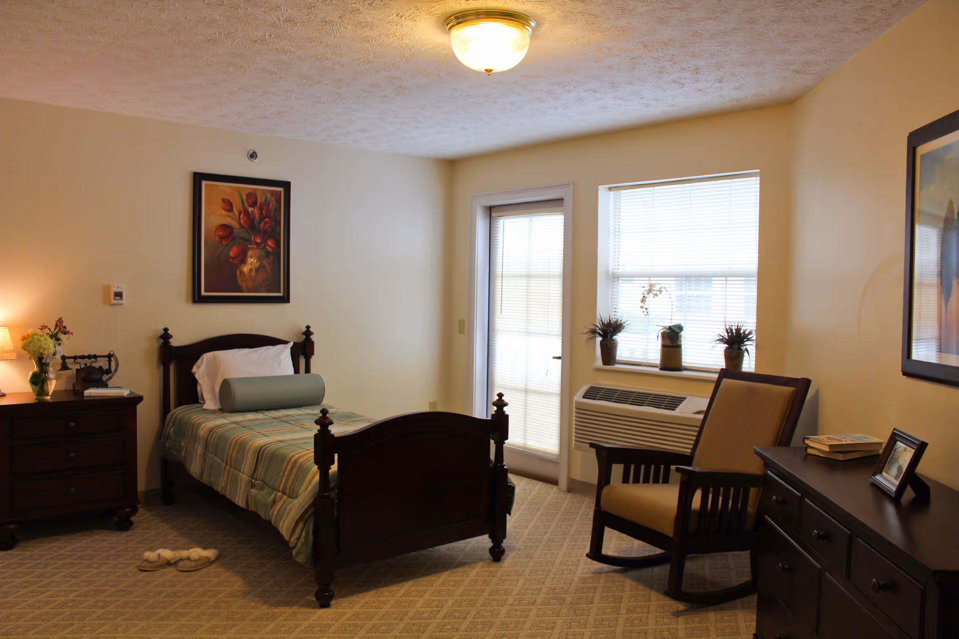 A cozy bedroom with a single wooden bed covered in a striped bedspread and a bolster pillow. There is a nightstand with a lamp, flowers, and a vintage telephone on the left side of the bed. A framed painting of flowers hangs above the bed. To the right, there is a window with blinds and a door with glass panels. Below the window is an air conditioning unit with three small potted plants on the windowsill. A wooden rocking chair is positioned next to a dresser with books and a framed photo on top. The room has beige walls and carpeted floor with a subtle pattern.