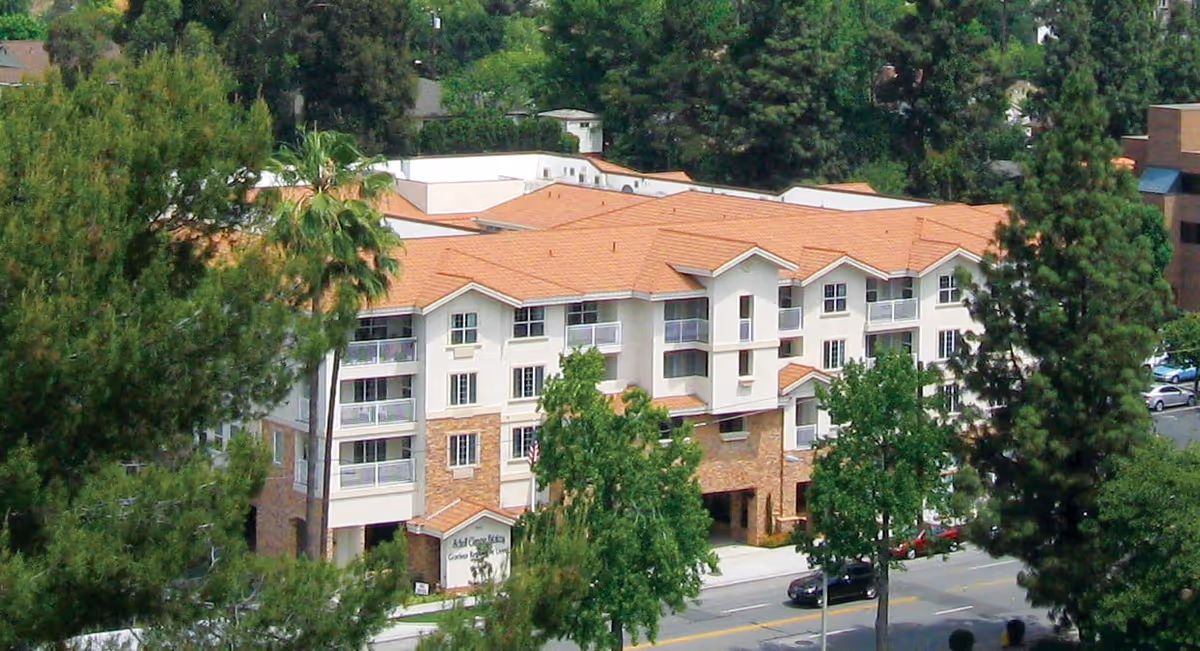 A multi-story residential building with a tiled roof surrounded by trees and greenery, viewed from an elevated angle. The building has balconies and large windows, with a street and parked cars visible in front.