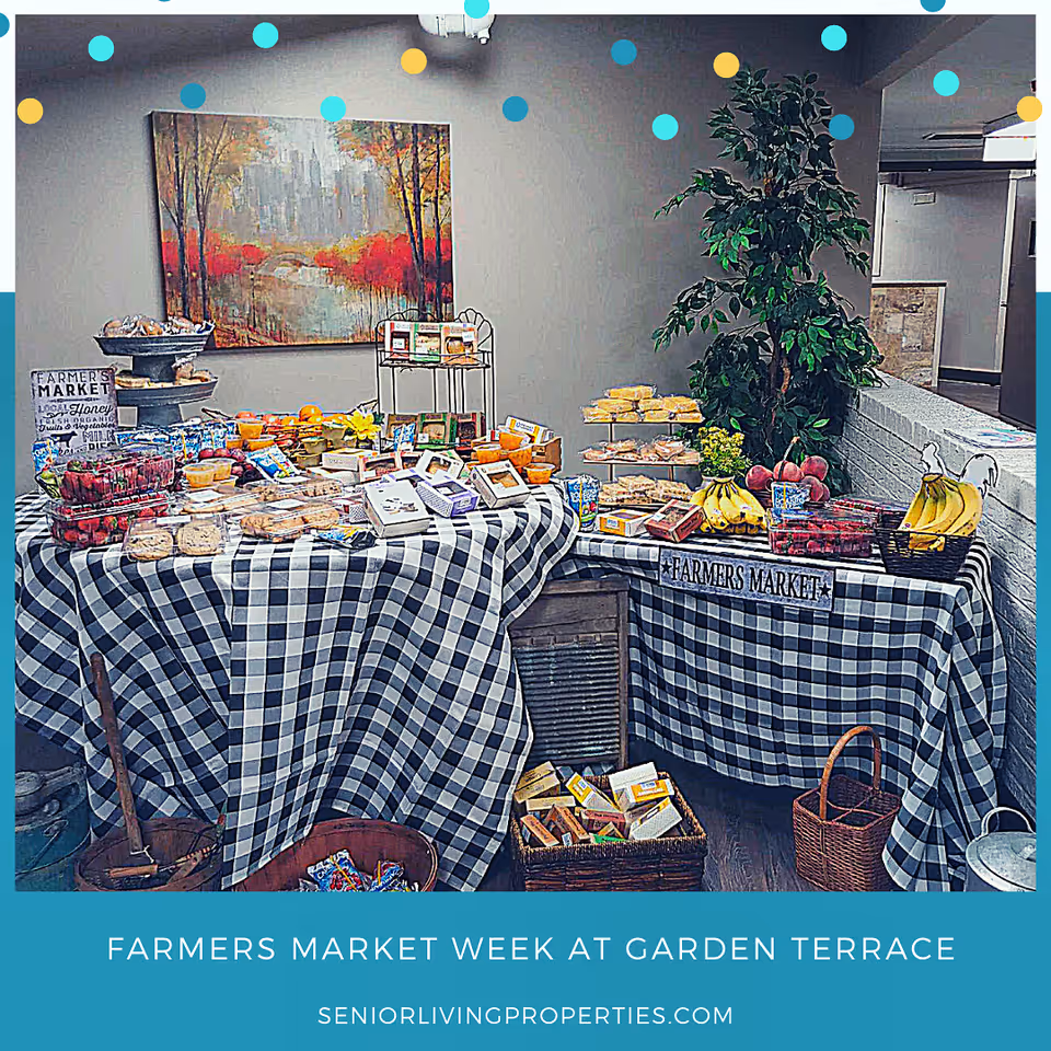 Indoor farmers market setup with tables covered in black and white checkered tablecloths displaying various fruits, packaged snacks, and baked goods. There is a large painting of a scenic autumn landscape on the wall behind the tables and a tall green plant to the right. The setting appears to be inside a senior living facility.