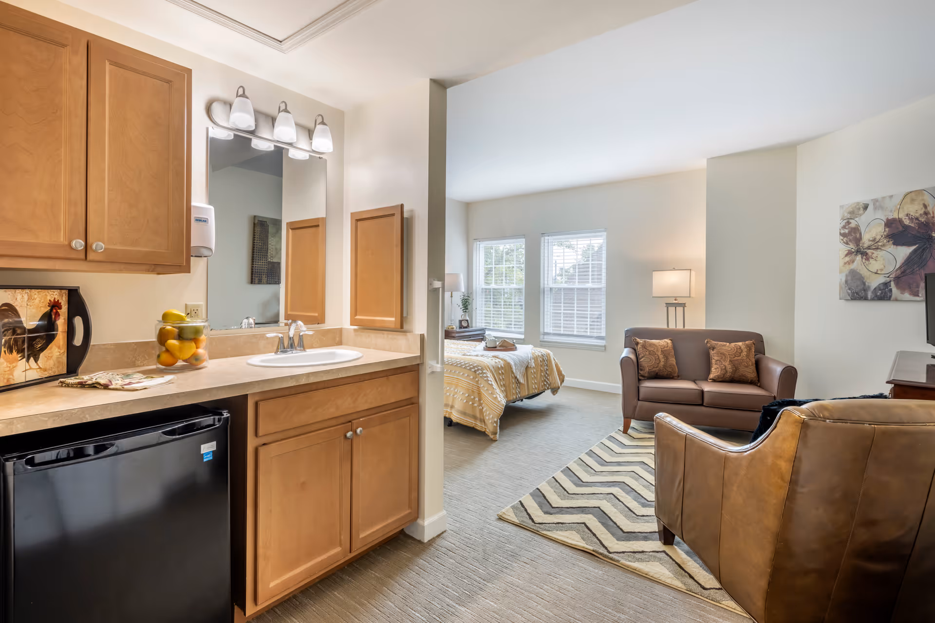 A bright and cozy studio apartment featuring a small kitchenette with wooden cabinets, a sink, and a mini refrigerator. The living area includes a brown leather armchair, a small brown loveseat with two patterned pillows, a chevron-patterned rug, and a bed with a yellow and white patterned blanket near two windows with white blinds. A floor lamp and a piece of floral wall art are also visible.