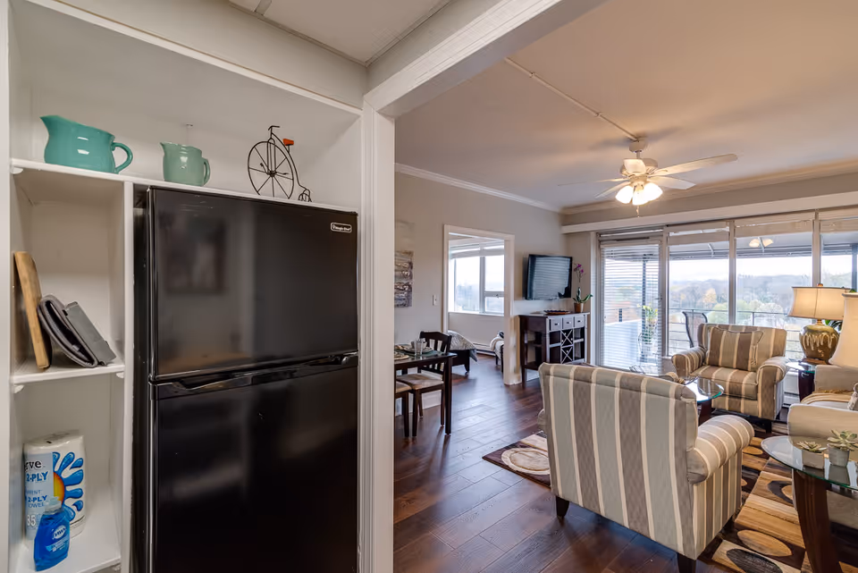 View of a cozy apartment interior showing a black refrigerator in a small kitchen nook with shelves holding decorative pitchers and paper towels. The image opens into a living room with striped armchairs, a sofa, a glass coffee table, a ceiling fan with lights, a wall-mounted TV, and large windows with blinds overlooking an outdoor balcony and scenic landscape.