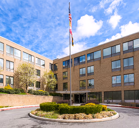 Exterior view of a three-story brick building with multiple windows, a circular driveway with a landscaped island in the center, and three flags on a tall flagpole against a partly cloudy blue sky.
