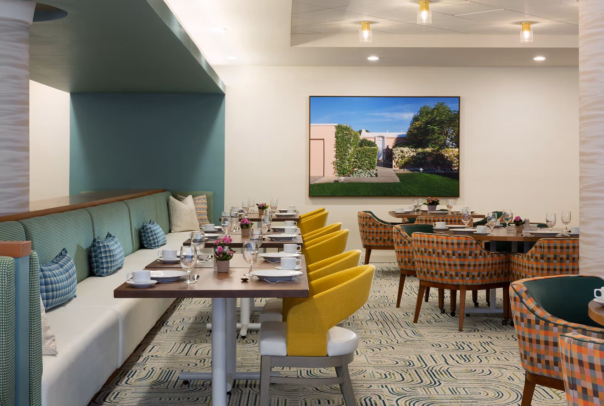 A modern dining room with a long table set for a meal, featuring yellow and patterned chairs. The room has a cushioned bench along one wall with blue and white pillows. A large framed photograph of a garden scene hangs on the wall, and the floor is covered with a patterned carpet.